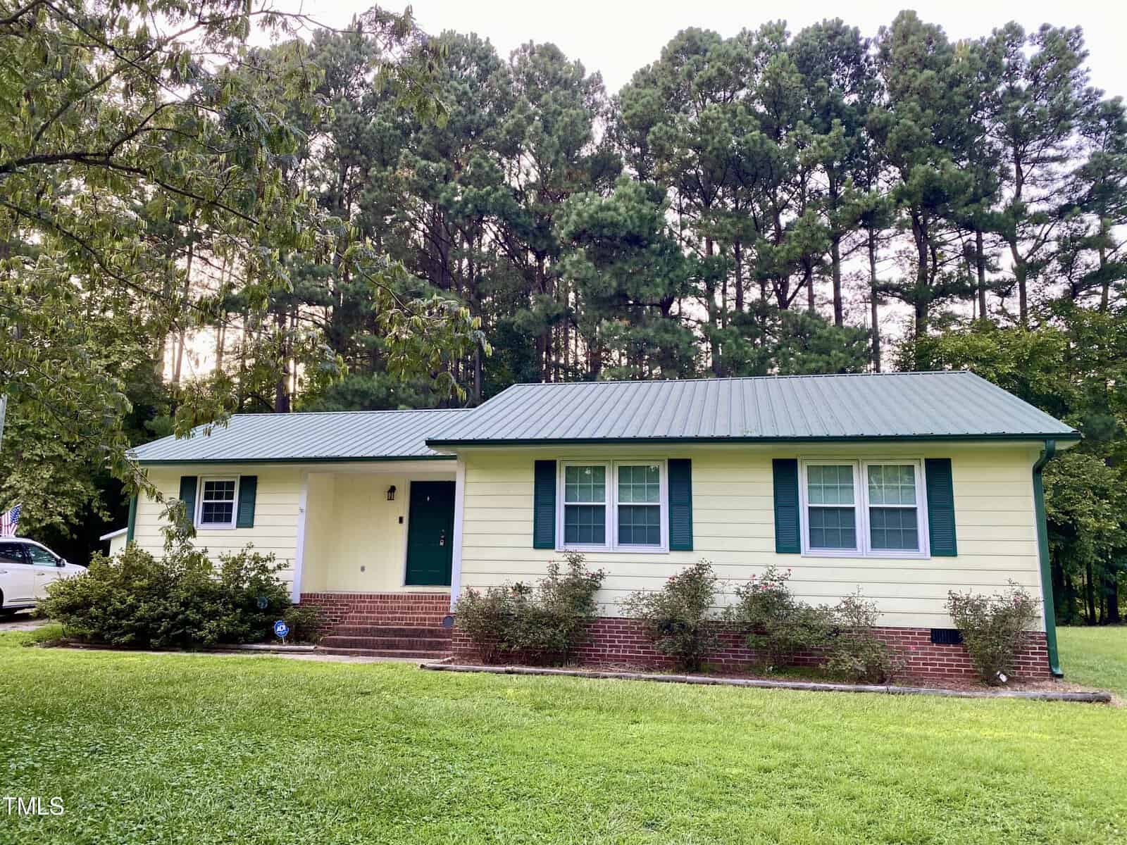 A single-story light yellow house with a green metal roof and green shutters, set against a backdrop of tall trees, located at 3240 US 158 Bypass, Henderson. The house has white trim and a front porch with brick steps leading up to a green front door. There is a well-maintained lawn with shrubs.