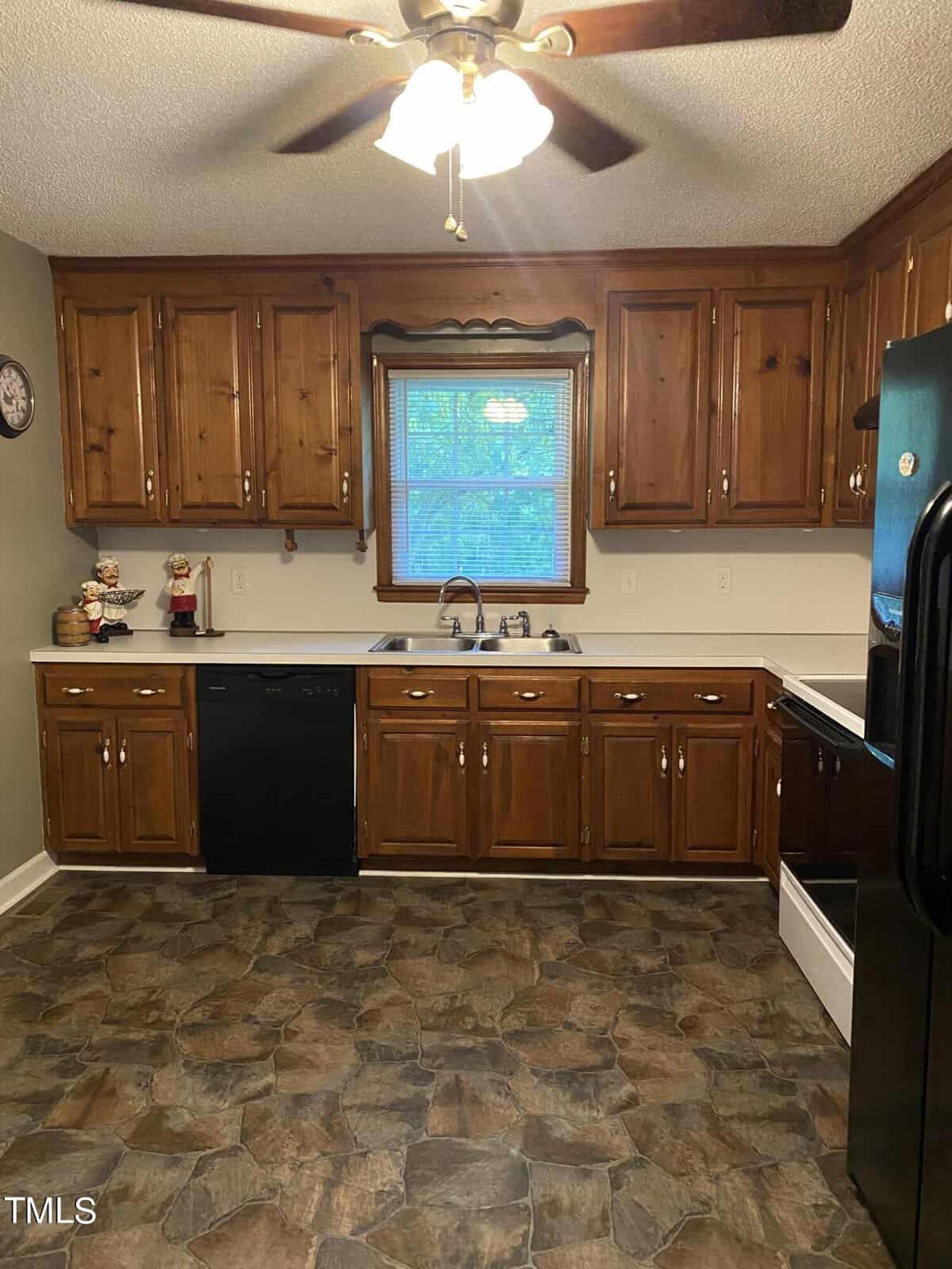 A kitchen featuring wooden cabinets, a double sink under a window, and a black dishwasher to the left. The floor is covered in a stone-patterned vinyl. An overhead ceiling fan with lights is visible. The walls have a clock and a small decorative figurine.