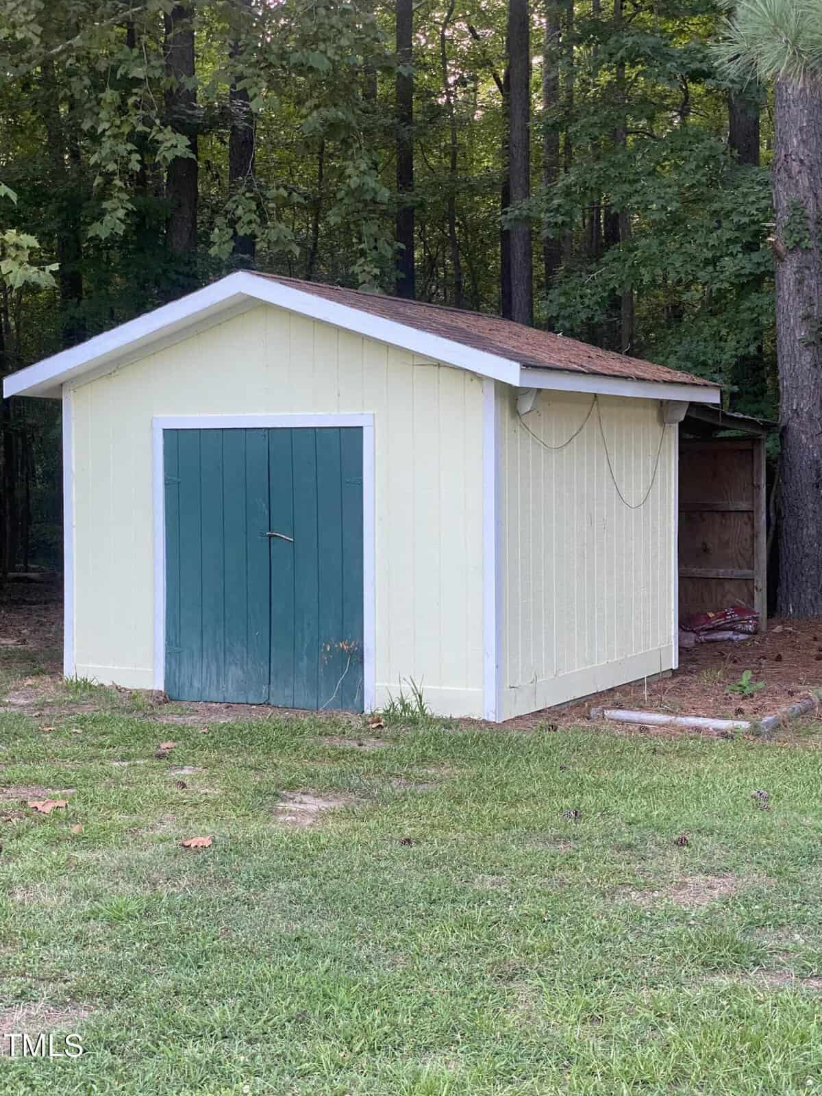 A small, pale yellow shed with a sloped roof and a green double door stands on a grassy area surrounded by trees. A coil of wire hangs from the roof's edge. The shed is adjacent to a wooded area, providing shade and a natural backdrop.
