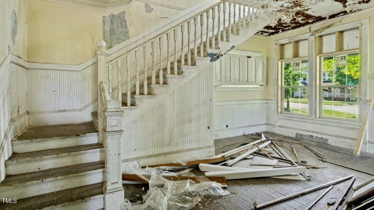 A dilapidated interior of a house featuring a wooden staircase with peeling white paint. The walls and ceiling are in disrepair, and debris, including broken wood and plastic sheets, litters the dark wooden floor. Sunlight streams in through the large windows.