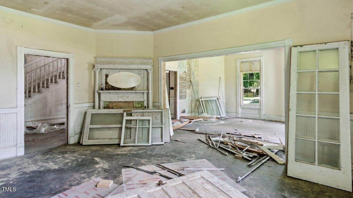 An abandoned room with peeling paint and scattered debris. There are several old window frames leaning against walls. A set of glass panel double doors is open, leading to another cluttered room. An old fireplace with a mirror above it is visible.