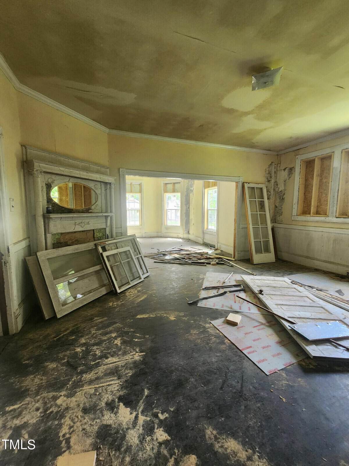A dilapidated room with damaged floors and walls, scattered debris, and broken windows leaning against a wall. The room has a fireplace with a missing mantle and a large doorway leading to a sunlit space with boarded-up windows. The ceiling shows signs of water damage.