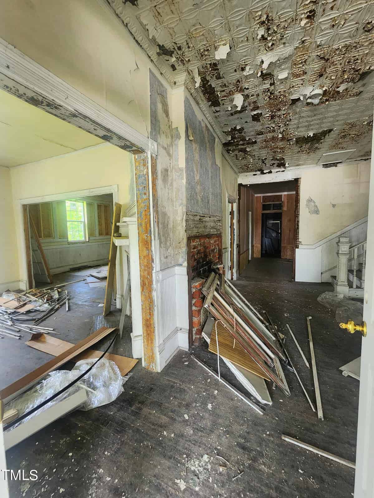 An interior view of an abandoned, dilapidated house with peeling paint, exposed brickwork, and scattered debris and materials on the floor. The ceiling shows significant water damage and decay, revealing worn plaster and exposed wood. Sunlight filters in through a window.
