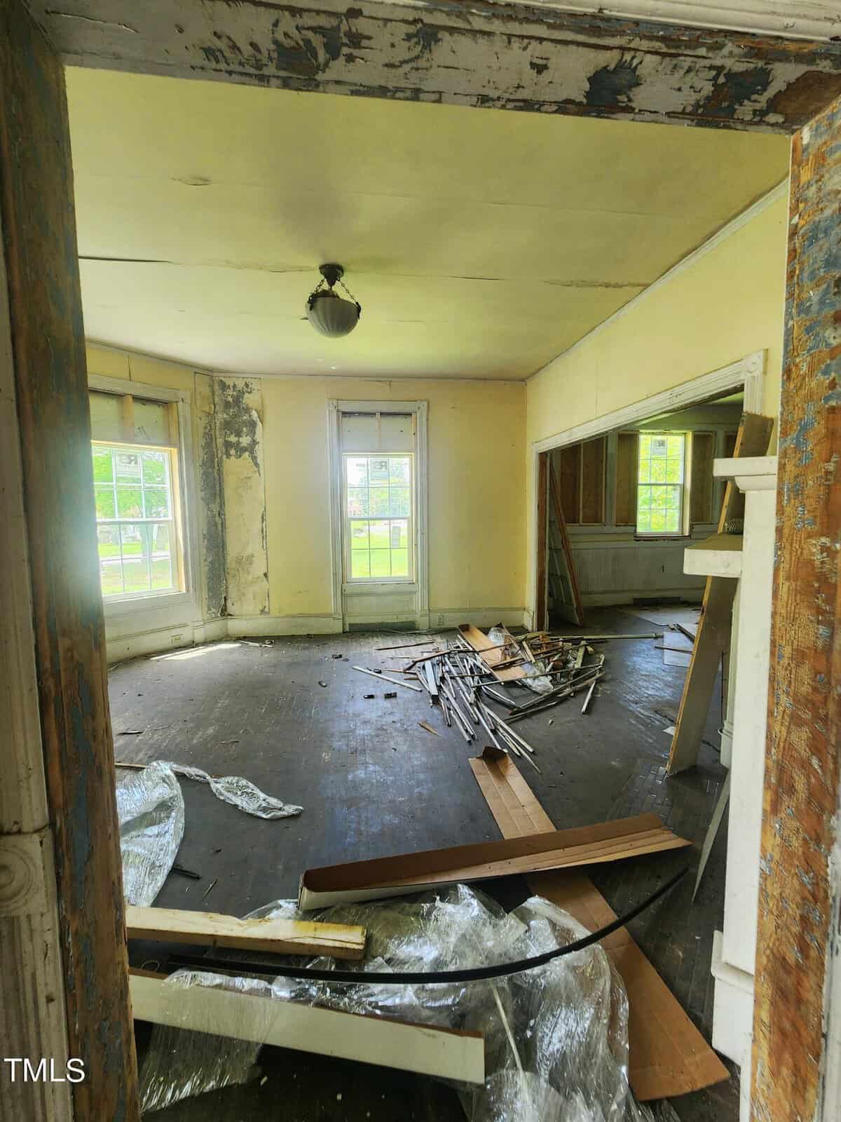A dilapidated room with wooden floors partially covered in debris. The walls are peeling, and a pile of dismantled wooden planks is in the center. Large windows allow natural light to enter. A light fixture hangs from the ceiling. The room has two doorways.