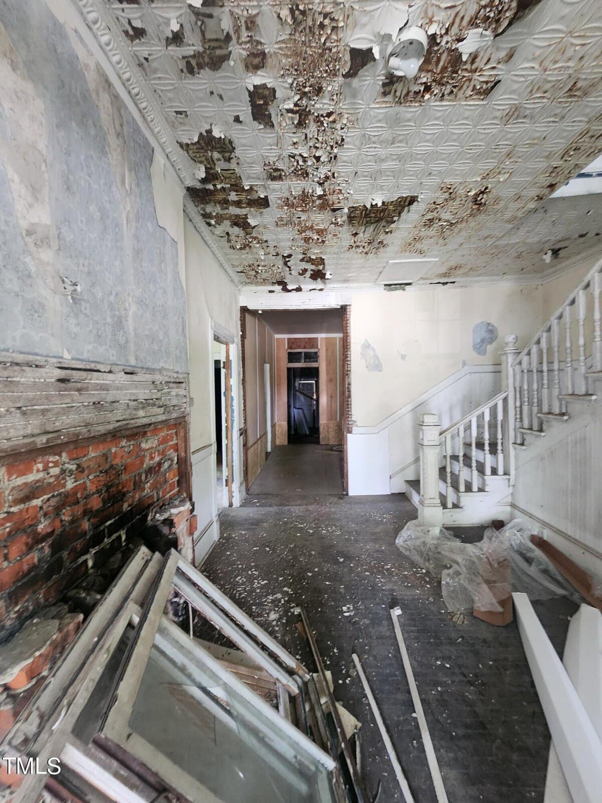 A dilapidated interior of a building showing extensive ceiling damage with peeling paint and exposed brick. The floor is cluttered with debris, including broken window frames. A staircase with white railings ascends on the right side of the image.