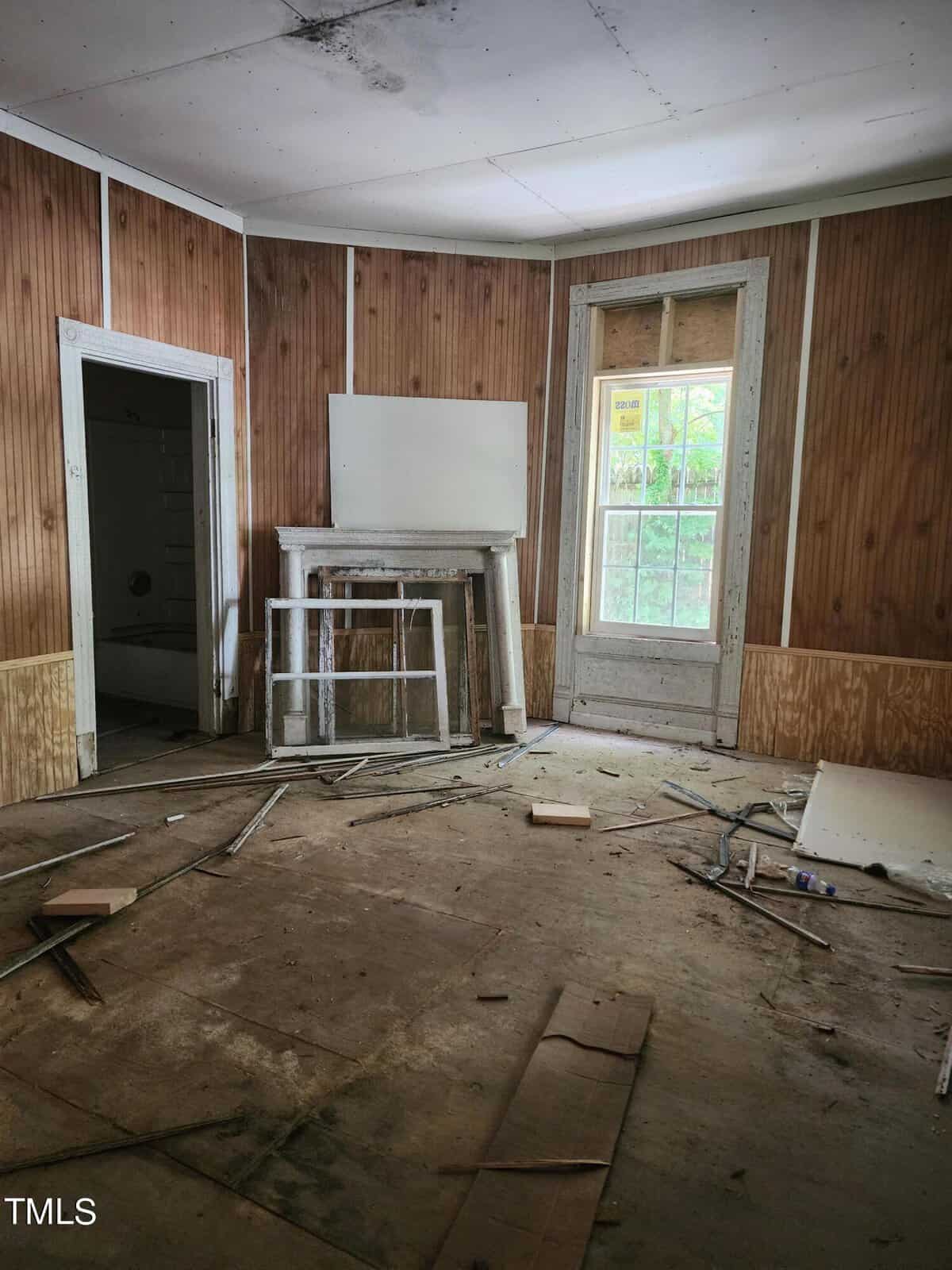 An abandoned room with wood-paneled walls, a partially open door, and a window with a "Beware" sign. The floor is cluttered with debris, broken wood, and scattered metal rods. A white mantlepiece is in front of the window, and the ceiling shows signs of damage.