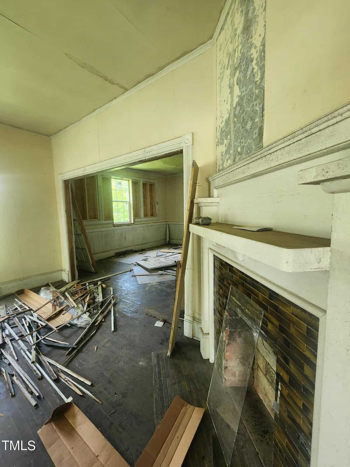 A dilapidated room filled with scattered debris, wooden planks, and broken glass. The walls have peeling paint and exposed wall studs. A non-functional fireplace with a weathered mantel is on the right. Sunlight filters through a dirty window in the background.