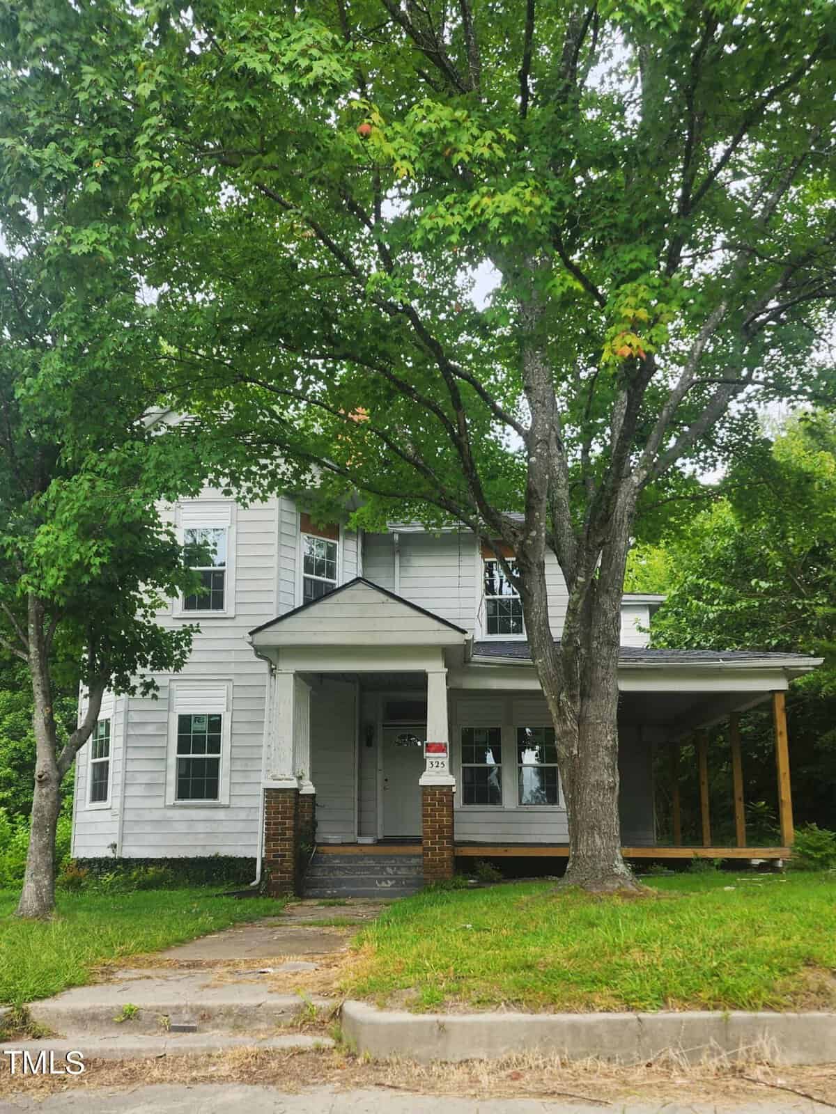 A white two-story house at 325 Charles Street features a small front porch and steps leading to the front door. Partially obscured by large trees, the Henderson property boasts a grassy yard with a concrete path. A "For Sale" sign stands prominently near the front steps.