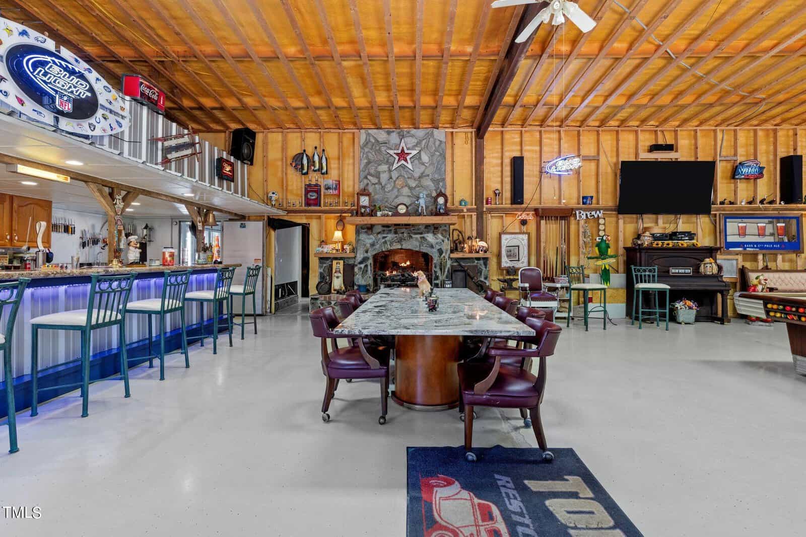 A spacious, rustic-themed bar area with a long marble table surrounded by chairs. The room features a wooden ceiling, a stone fireplace with a star above, several neon signs, a bar counter with stools, and vintage decor items.