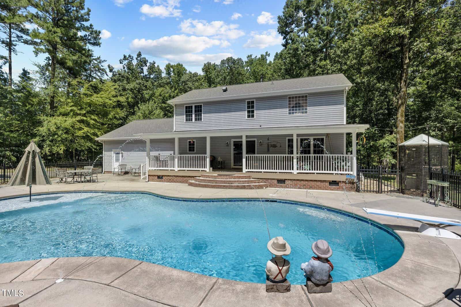 A spacious two-story house with a large backyard featuring a swimming pool. Two decorative figures resembling children sit at the pool's edge. The area is surrounded by lush trees and a patio with furniture is visible under a covered porch.