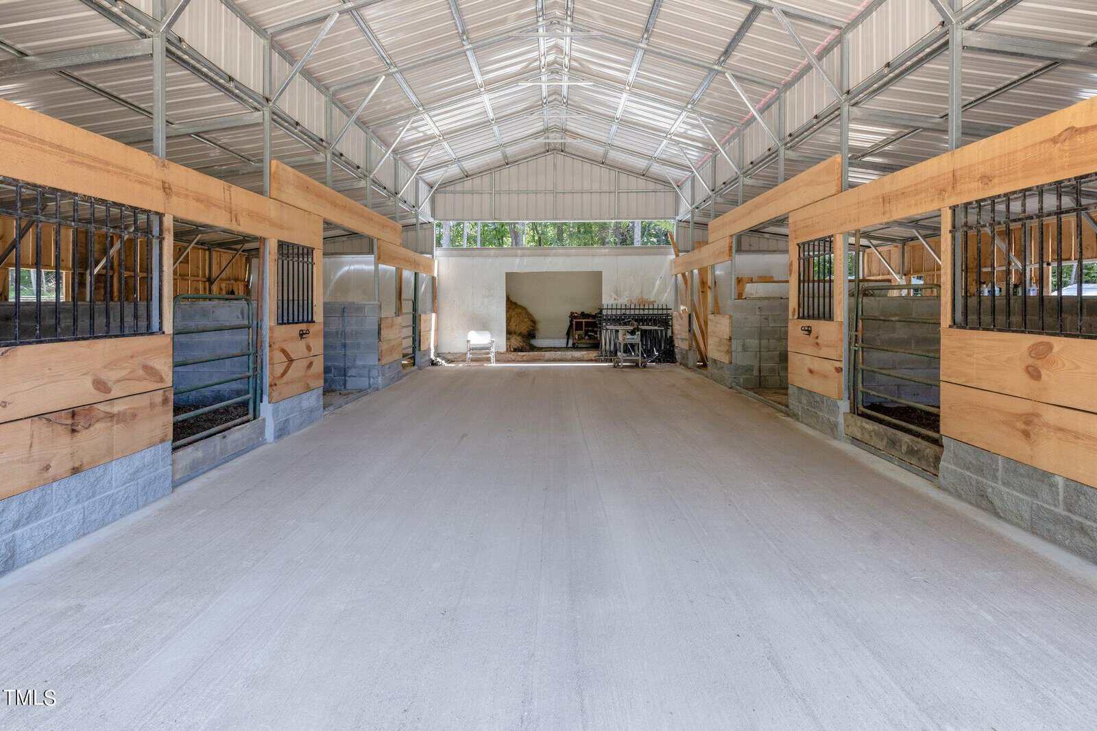 Interior of a modern horse stable with metal and wood construction. The stable features multiple stalls on both sides of a wide, concrete center aisle. Bright natural light comes in through the open doors at the far end.