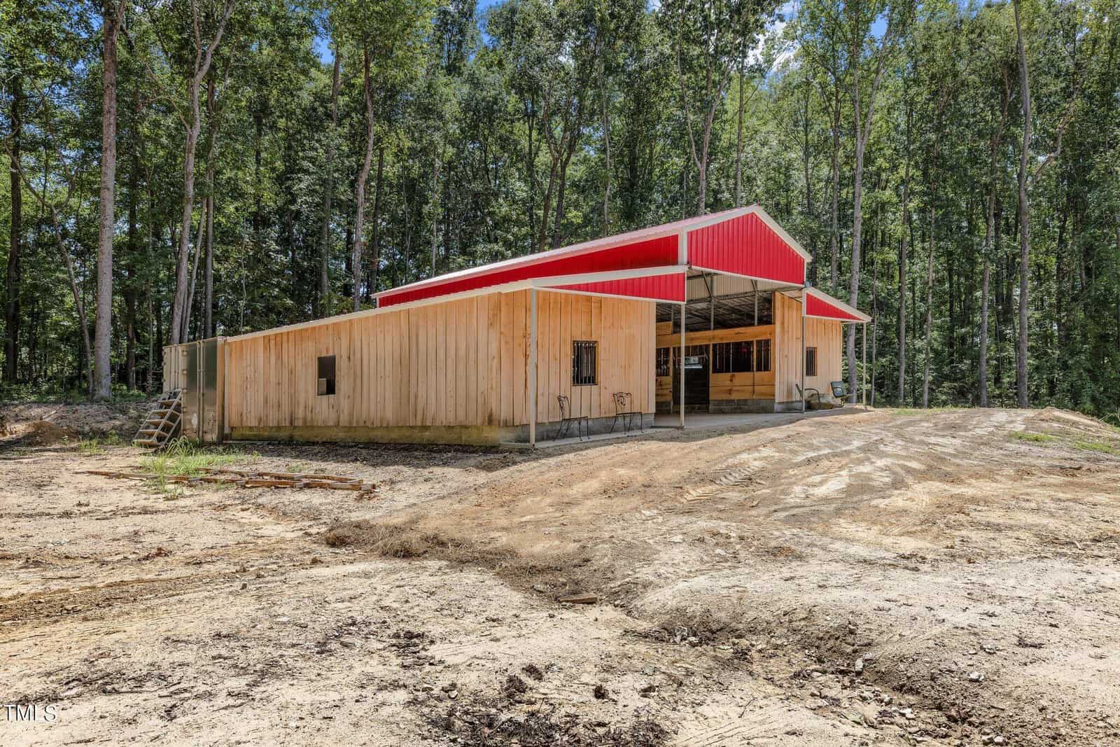 A large wooden barn with a red roof sits in a clearing surrounded by tall trees. The ground in front appears freshly cleared. The sky is clear, and the structure features multiple entry points and a spacious interior.