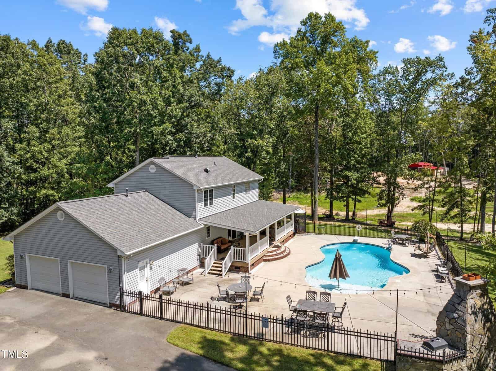 Aerial view of a two-story gray house with a spacious patio, outdoor seating, and a swimming pool. The property is surrounded by a black metal fence and lush trees, with a clear blue sky above.