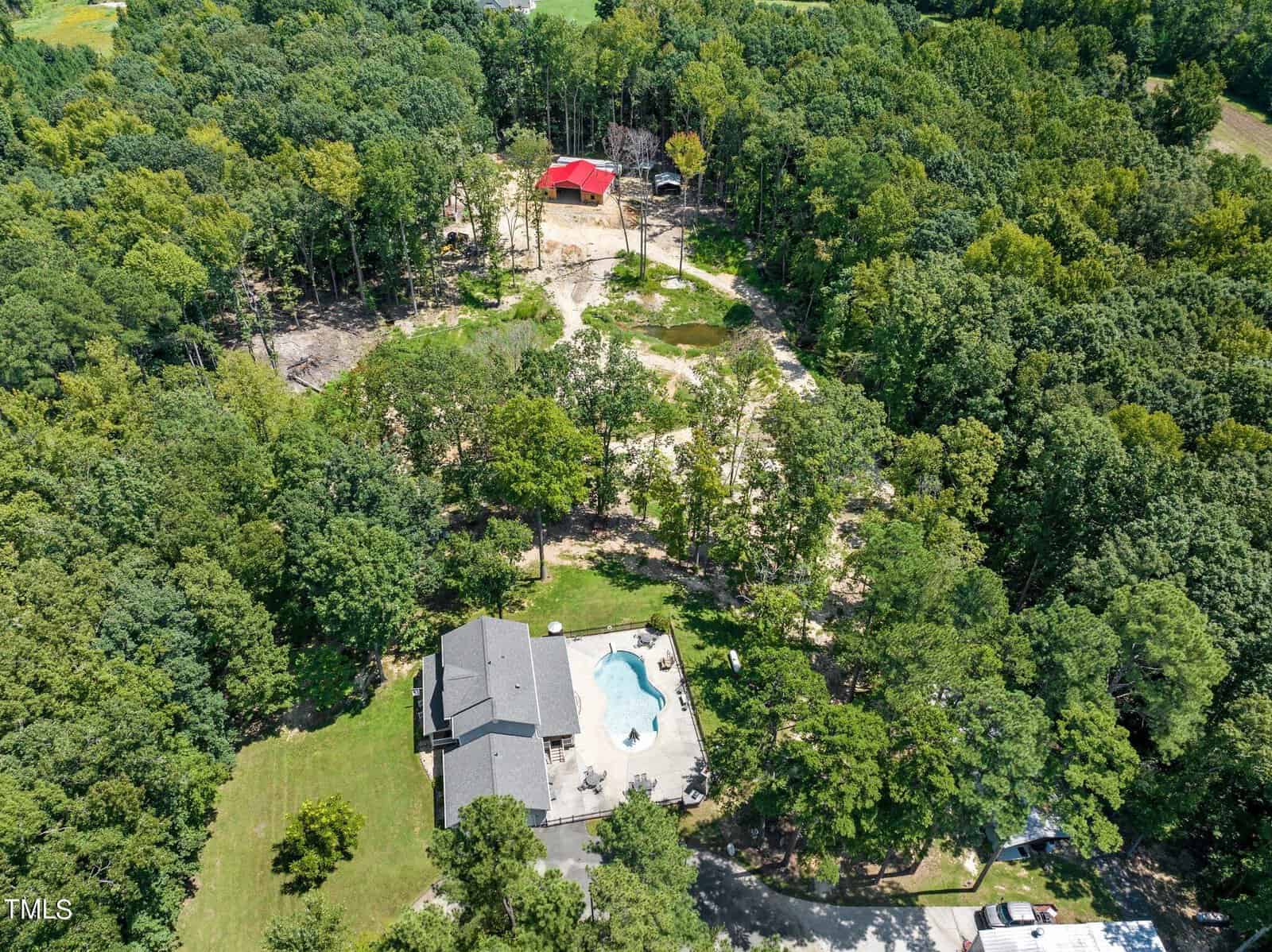 Aerial view of a wooded area with a large house and swimming pool surrounded by trees. There's a dirt path leading to a red-roofed building in the background. The scene is lush and green, indicating a rural setting.