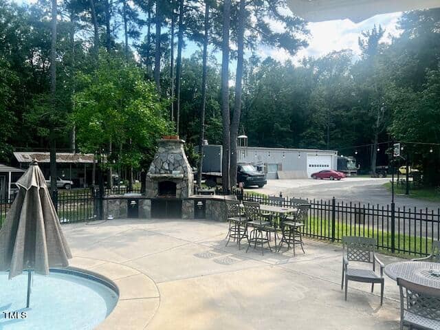 A backyard with a small pool, patio tables, and chairs surrounds a stone fireplace. A fenced area backs up to a driveway with a gray building, trees, and a truck parked nearby. The scene is sunny and peaceful.