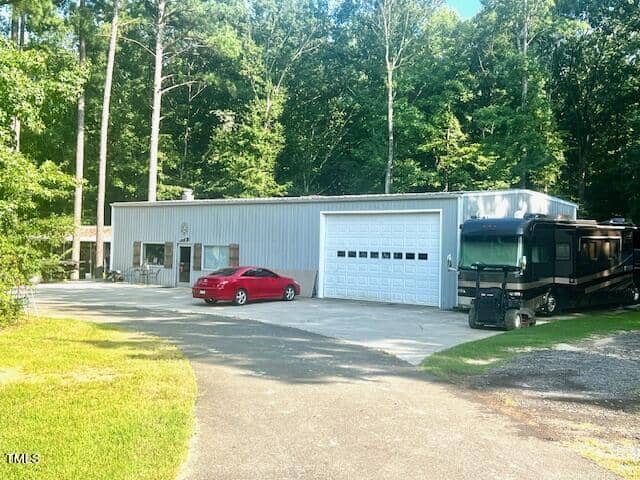 A large metal building with a garage door stands near a forested area. A red car is parked in front, and a black RV is parked to the side. The area is surrounded by tall trees and greenery.