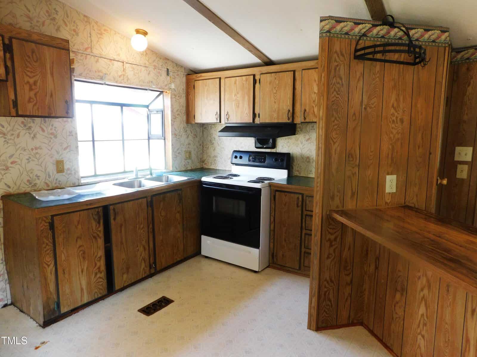 A rustic kitchen with wooden cabinets, floral wallpaper, and a white tile floor. It features a black and white electric stove, a double sink beneath a window, and wooden beams on the ceiling. There's a small built-in counter on one side.