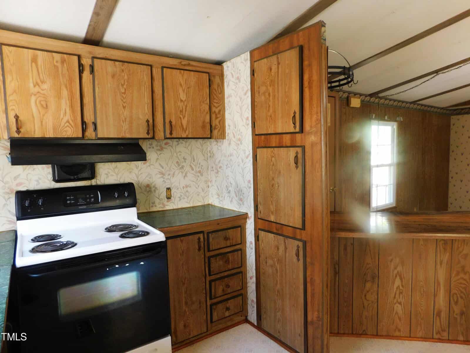 A kitchen with wood-paneled cabinets and light floral wallpaper. There's a white electric stove with a black hood and green countertops. A window is visible in the adjacent room, partially obscured by a narrow partition.