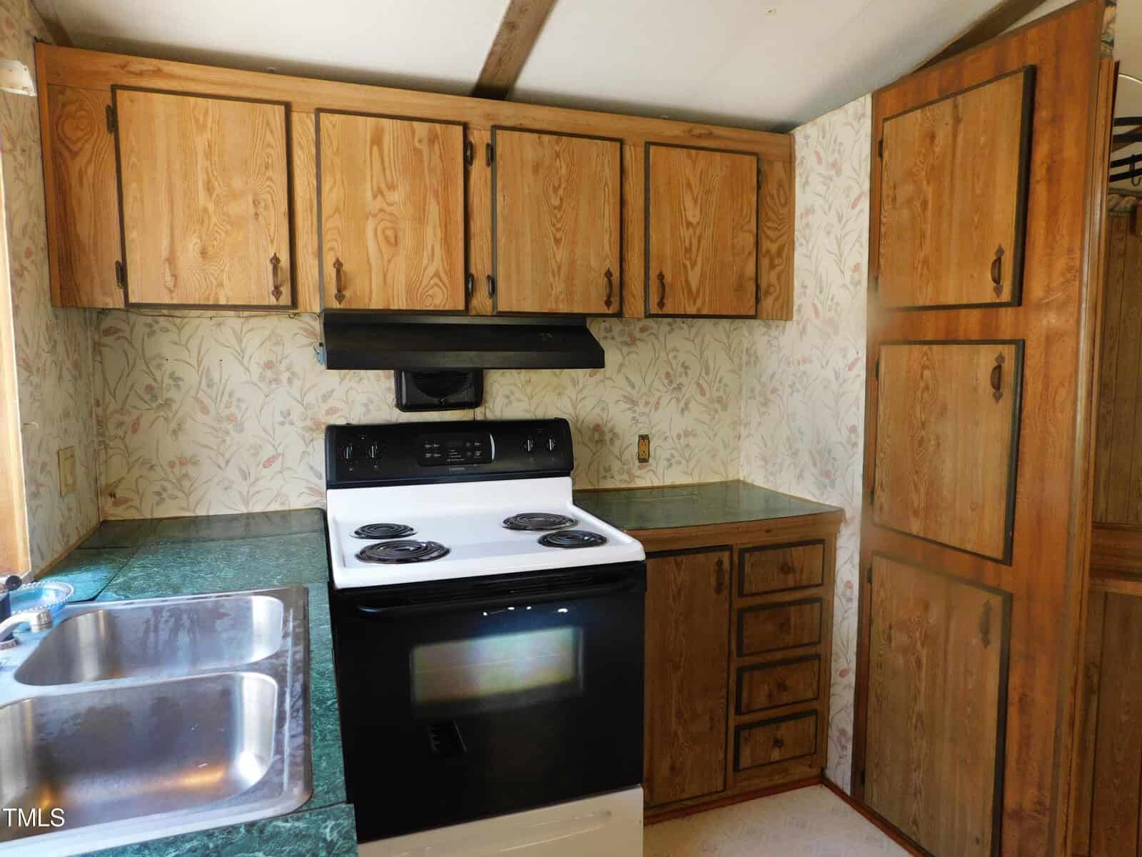 A compact kitchen with wooden cabinets and floral wallpaper. Features a white electric stove with black burners and a black range hood. Green countertops surround a double stainless steel sink. There's a tall cabinet on the right side.