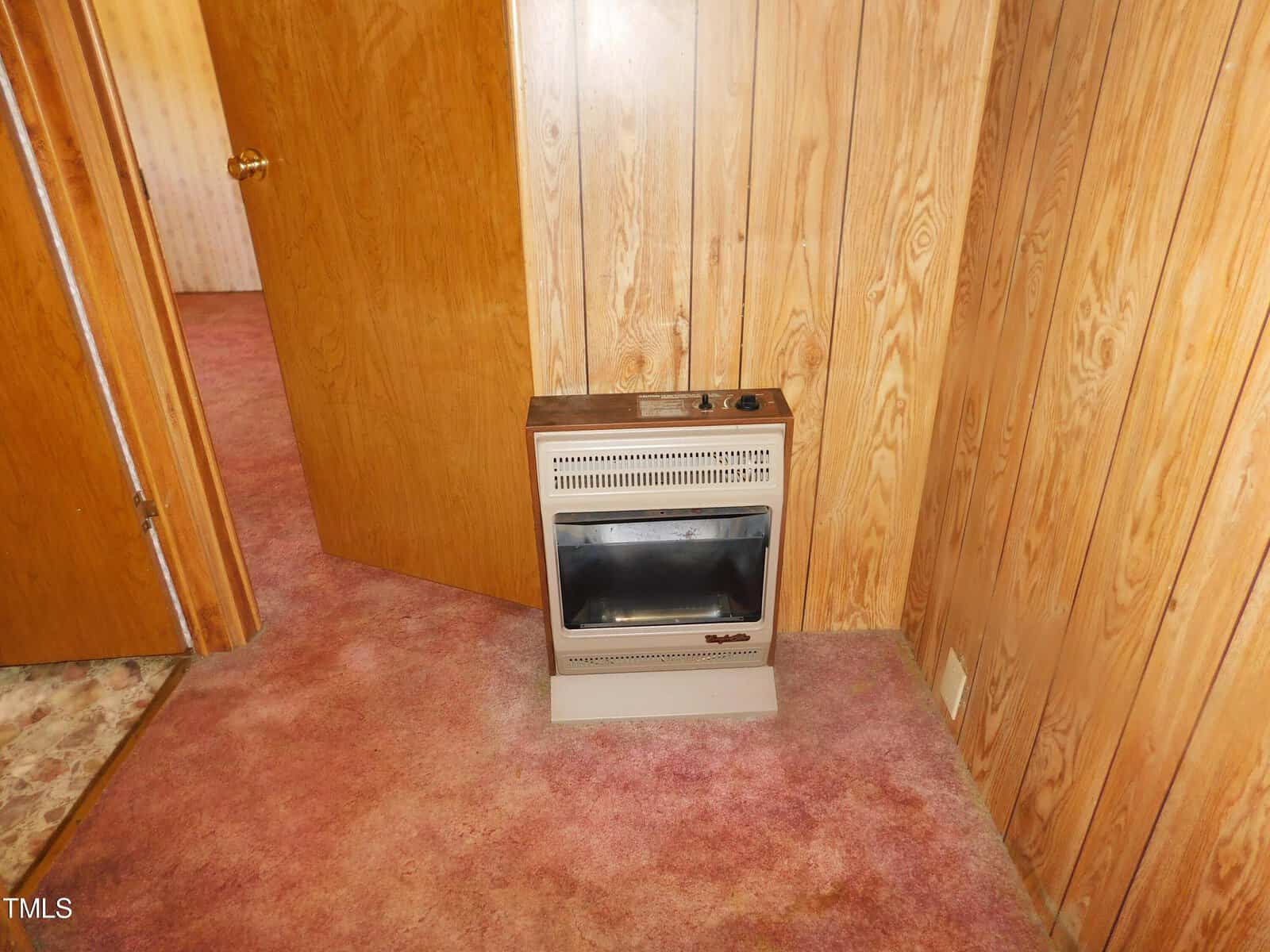 A small, vintage wall-mounted heater sits on a carpeted floor against wood-paneled walls. A partially open wooden door is visible on the left. The carpet is a faded reddish hue, adding to the retro aesthetic.