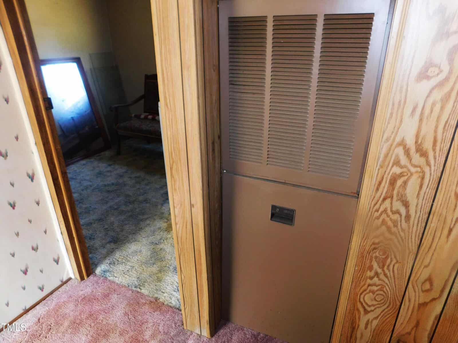 A brown HVAC unit is installed in a wooden wall between two rooms with different carpet colors. A chair and a framed picture are visible in the room on the left.