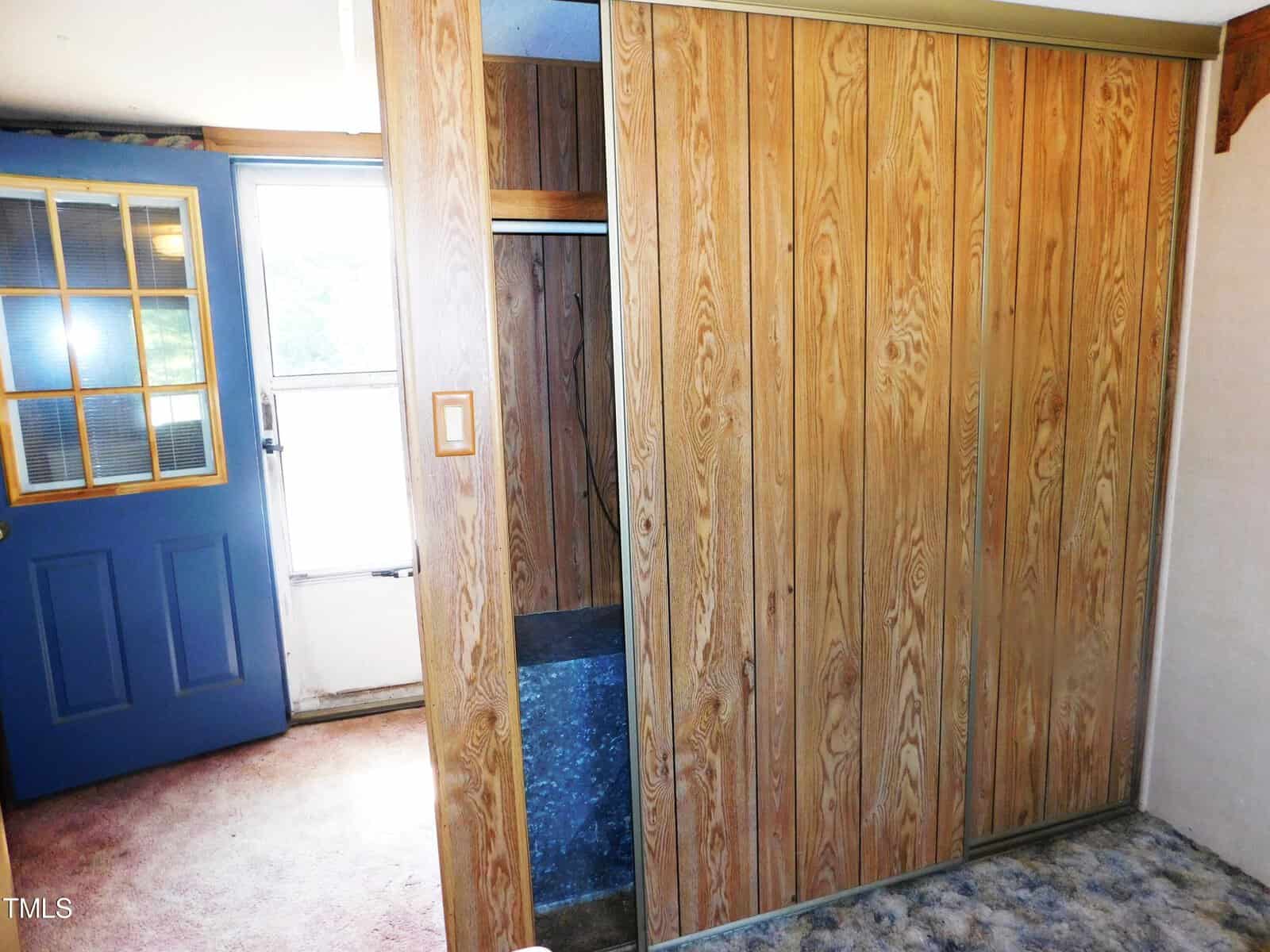 A small room with wood-paneled walls, a blue door with glass panes on the left, and a partially open closet with blue speckled carpeting. The room has a soft carpeted floor and a window letting in natural light.