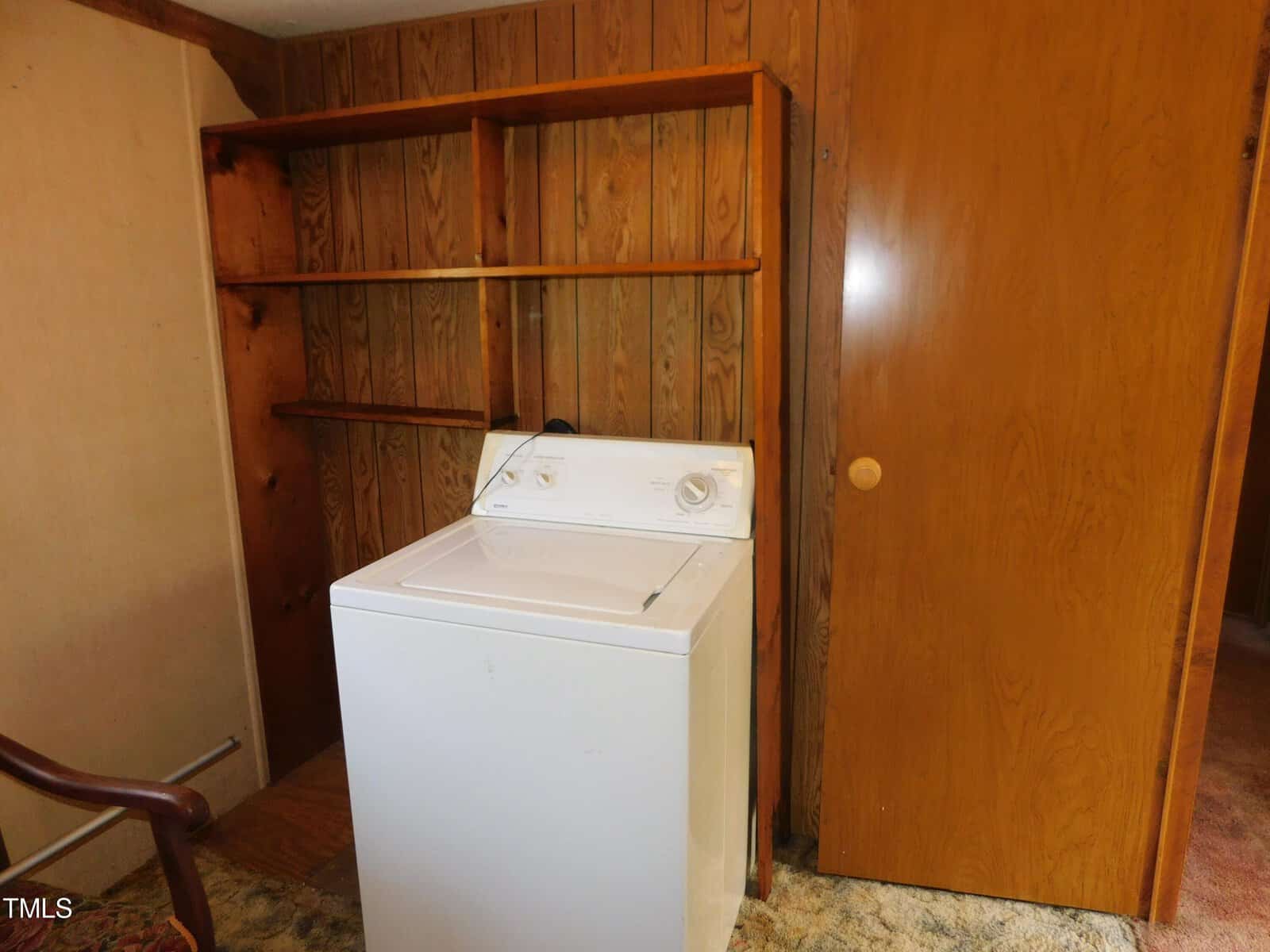 Laundry room with a white washing machine, wooden shelves, and wood-paneled walls. A wooden door is partially visible on the right. A chair's armrest appears in the lower left corner.