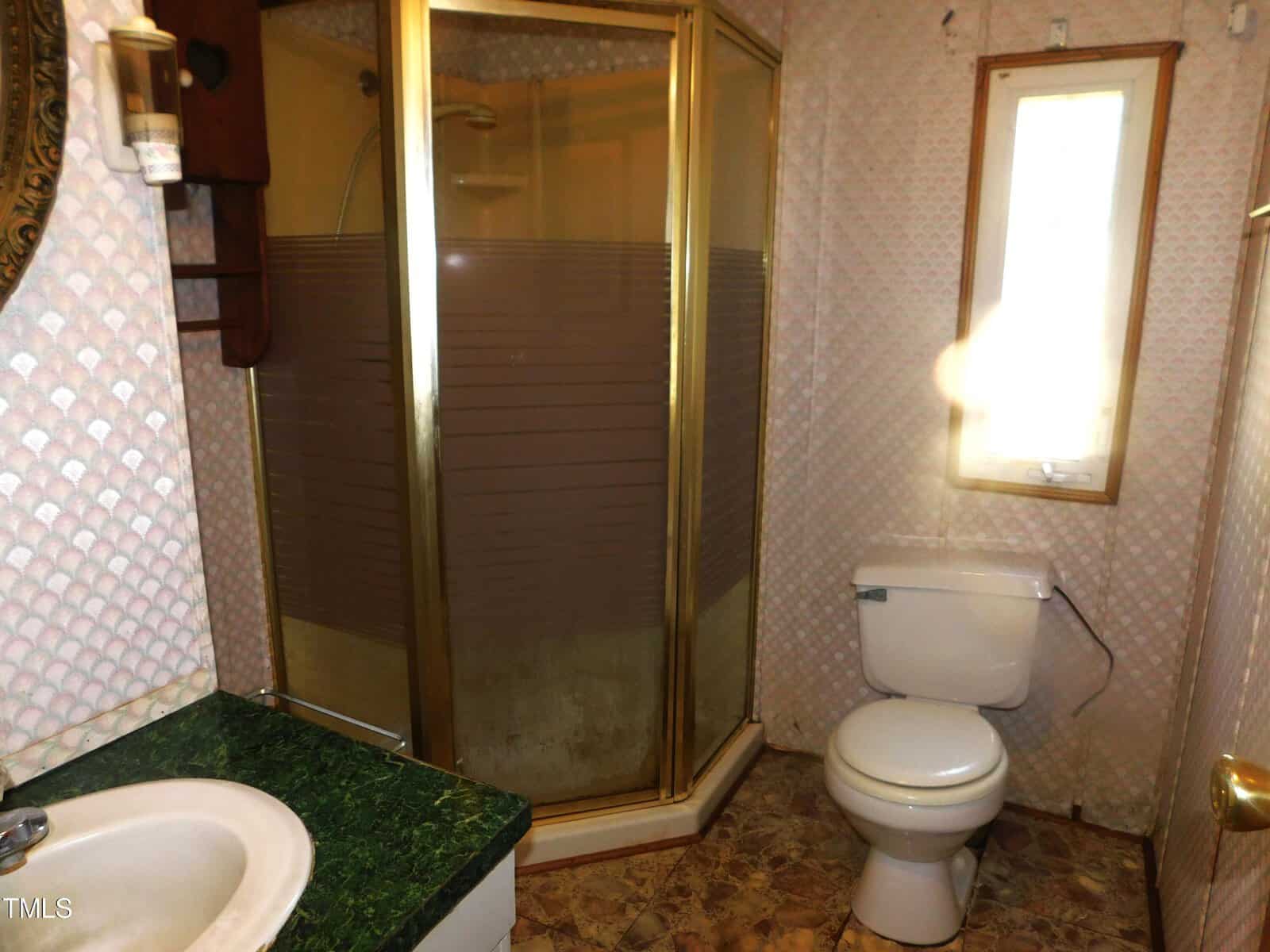 A small bathroom with a corner shower enclosed by glass doors, a toilet, and a sink with a green countertop. The walls are covered in patterned wallpaper, and a narrow window lets in natural light.