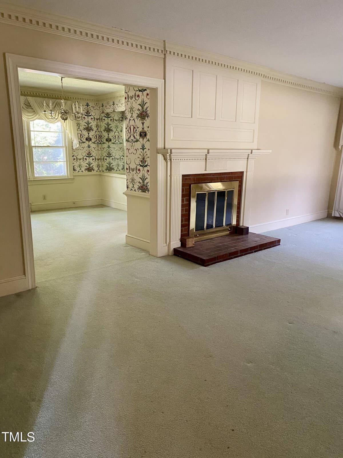 A spacious living room with light-colored carpet and a white, paneled fireplace with a brick base. An adjacent room with floral wallpaper and a window letting in natural light is visible. The walls and molding are painted in neutral tones.