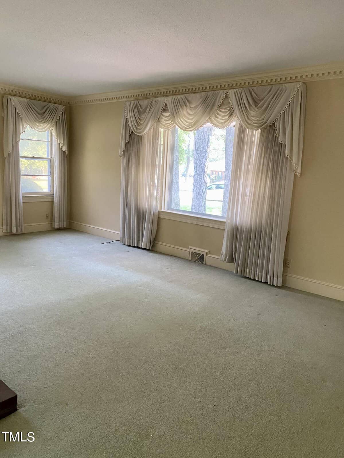 A spacious living room with beige walls and light green carpet. Large windows are adorned with elegant cream drapes, allowing natural light to flood the room.
