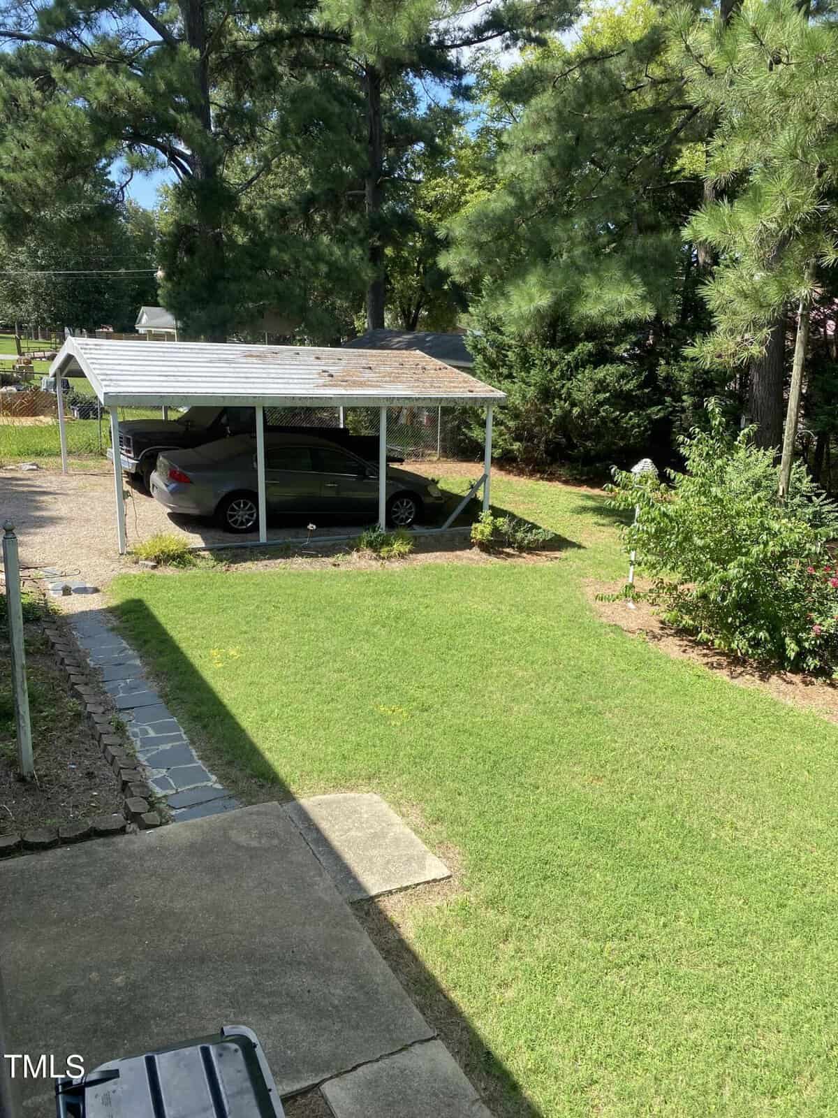 A backyard with a green lawn and a concrete path. A silver car is parked under a metal carport. Tall pine trees and shrubs surround the area, providing shade and privacy. A sunny day with clear skies.