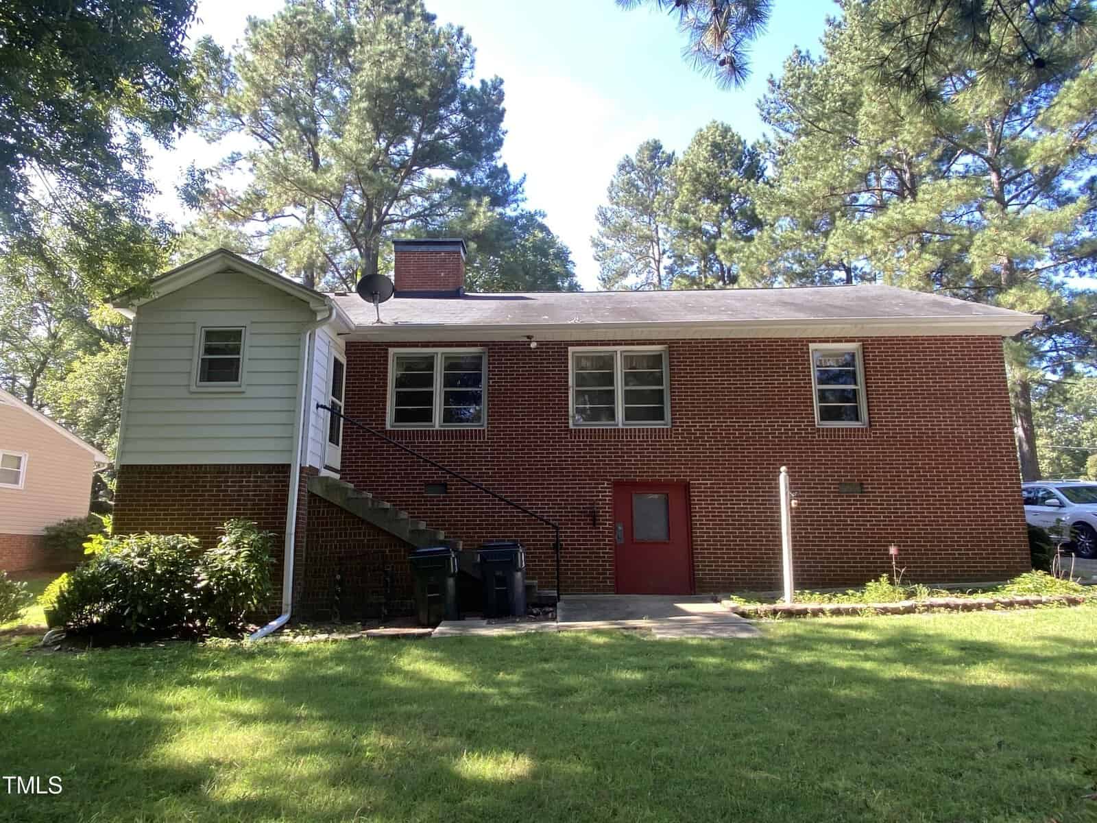 A brick house with a side staircase and red door, surrounded by a grassy yard and trees. The house has a chimney and several windows, set against a clear blue sky. Trash bins are placed near the stairs.