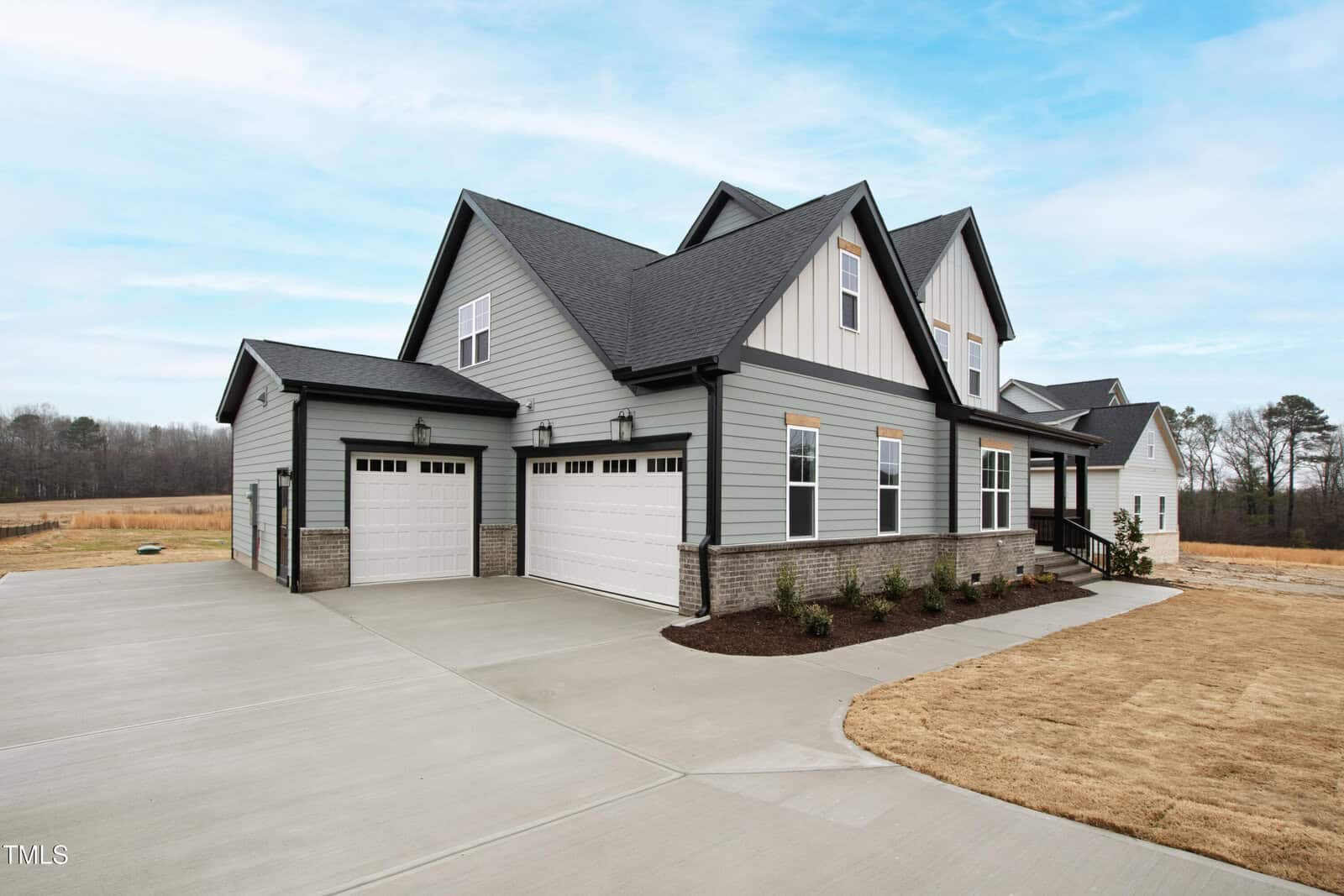 A modern two-story house with gray siding, white trim, and black roofing, featuring three garage doors and a large concrete driveway, set against a backdrop of open land and a blue sky.