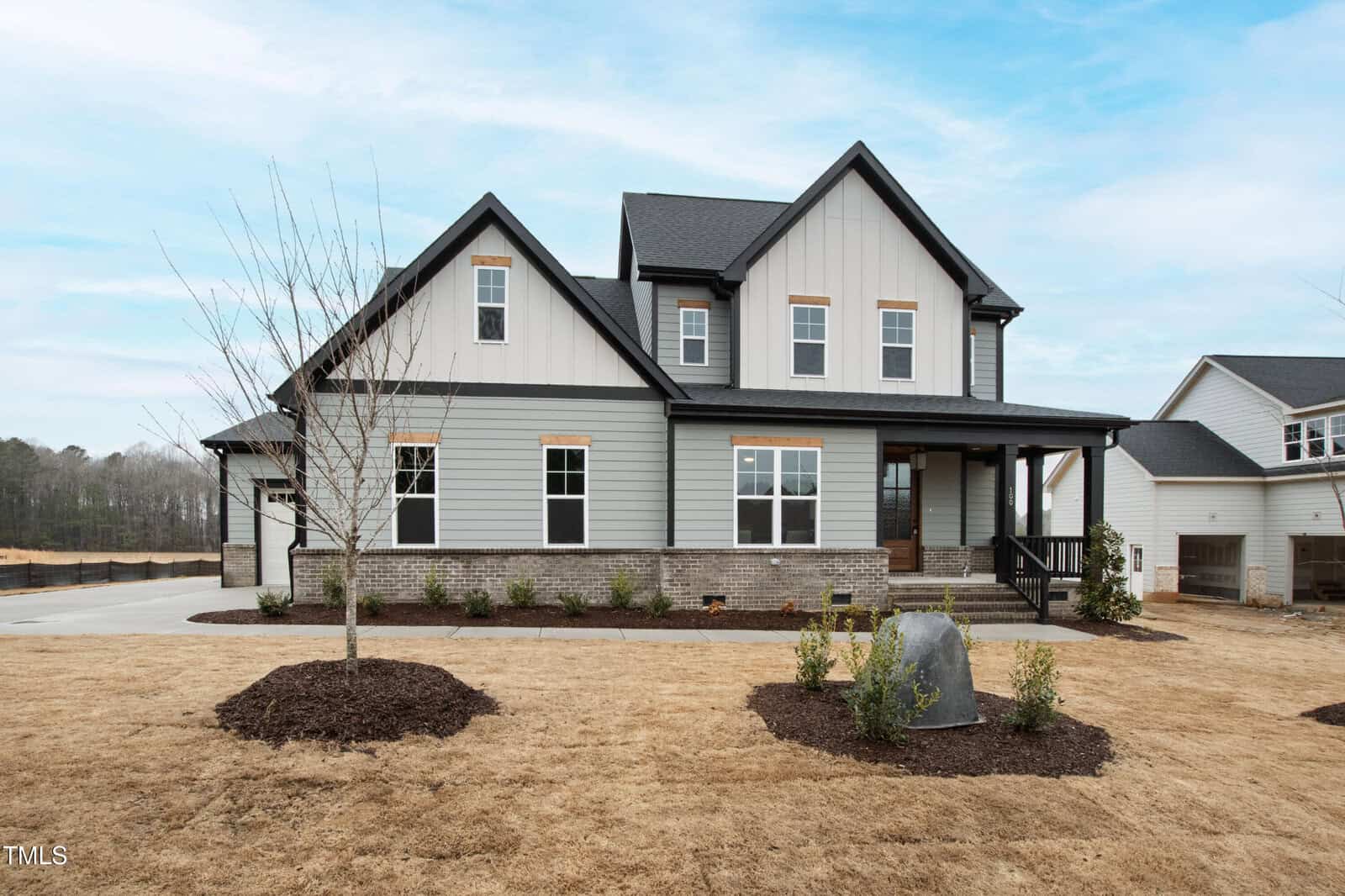Two-story house with gray siding, black roof, and a covered porch. The front yard has dry grass, small shrubs, a young leafless tree, and a large rock. A concrete driveway and neighboring houses are visible.