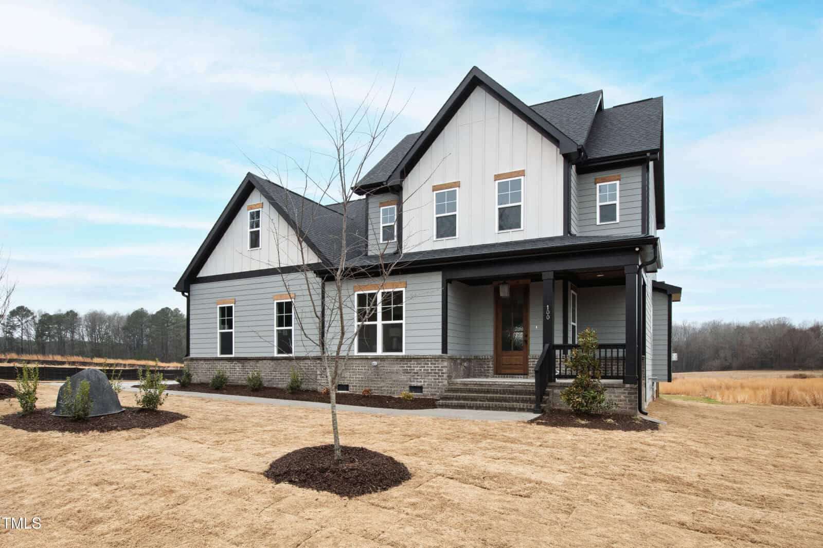 A two-story modern farmhouse with gray siding, black roof, large windows, and a covered front porch. The yard has fresh straw, young trees, and minimal landscaping, with open fields and trees in the background.