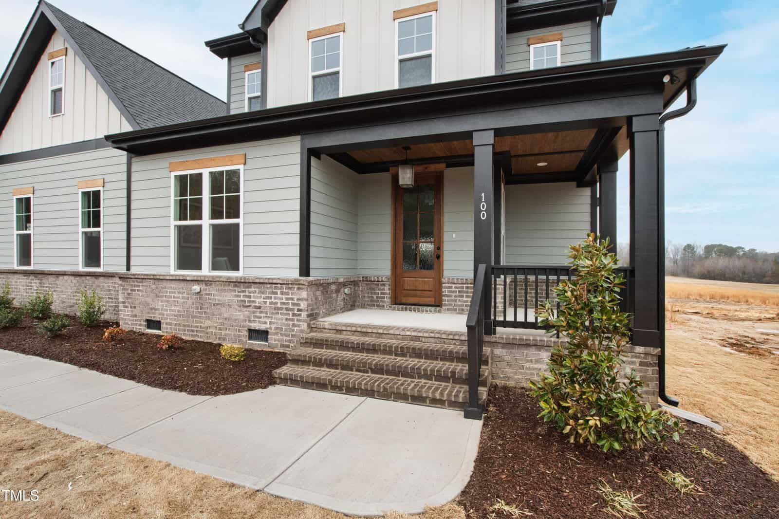 A modern two-story house with gray siding, brick foundation, white trim, and a covered front porch. The porch has black columns and railing, a wooden door, and a small landscaped garden along the sidewalk.