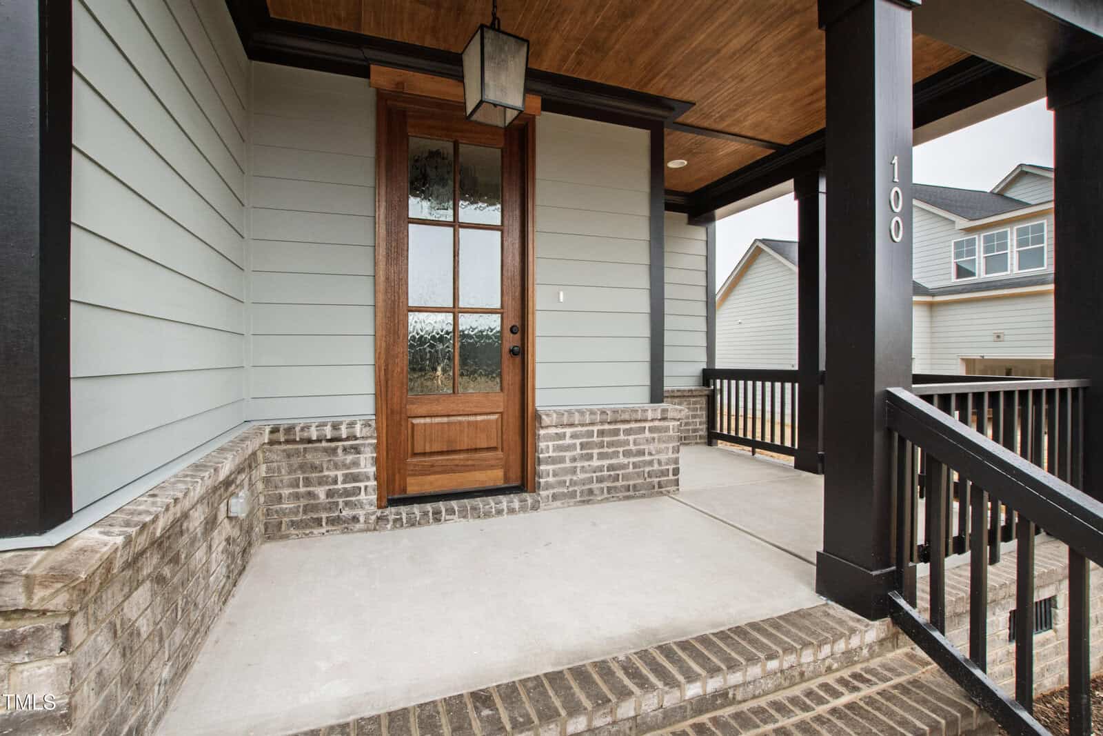 A modern front porch with a wooden door, frosted glass panels, brick steps, black railing, and gray siding, beneath a brown wood ceiling. House number 100 is visible on a black column.