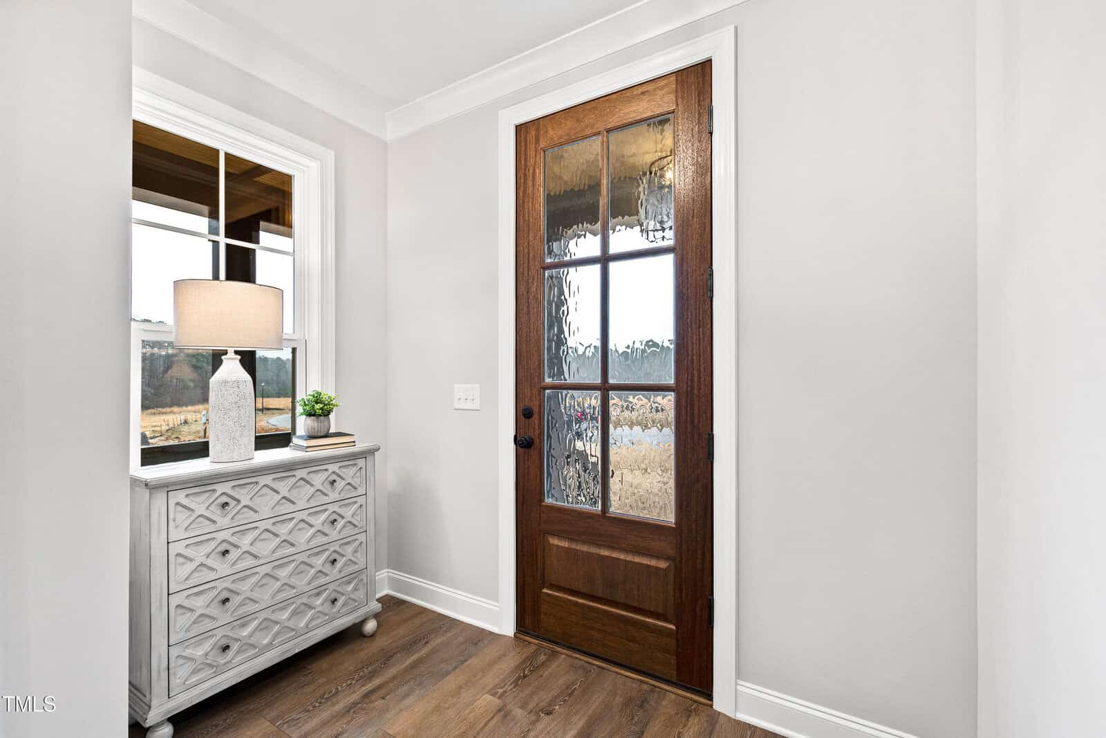 A wooden front door with decorative glass panels is next to a white console table with a lamp, a small plant, and books. The space has light-colored walls and wood flooring.