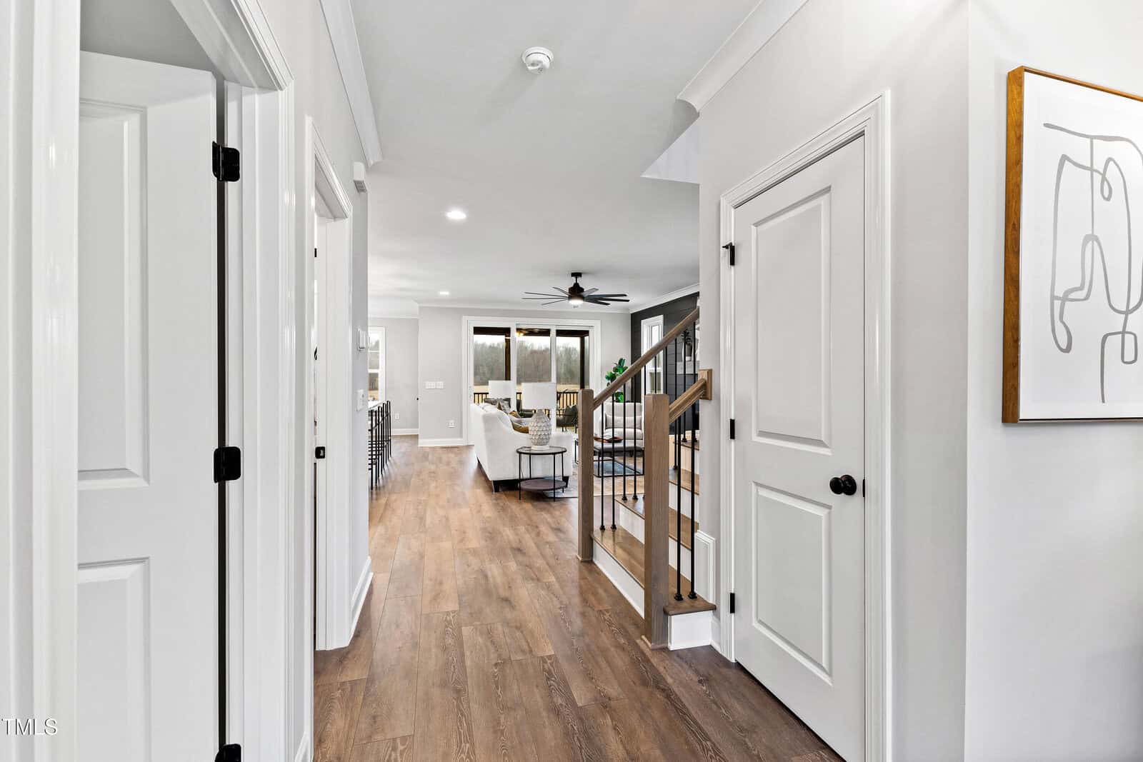 A modern hallway with light-colored walls and wood flooring leads to an open living area. The hallway has white doors, a staircase with metal railings, recessed lighting, and a framed abstract artwork on the wall.