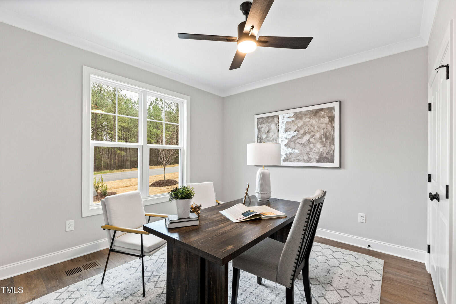 A modern home office with a dark wooden desk, three chairs, a table lamp, books, and a small plant. A large window lets in natural light, and there is a framed abstract artwork on the gray wall.