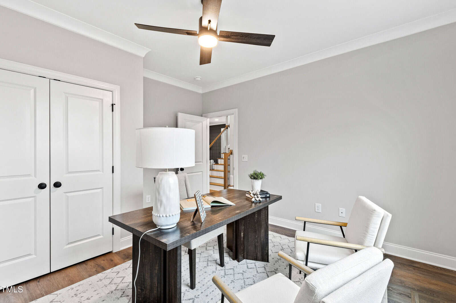 A modern home office with a dark wood desk, a white lamp, an open notebook, and two white chairs. Light gray walls, wood floors, a ceiling fan, and double doors are visible. An open door reveals a staircase.