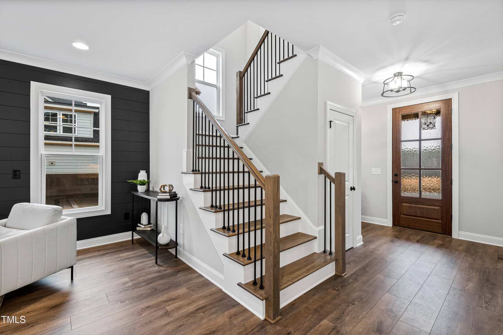 A modern entryway with wood flooring, a wooden front door with glass panels, an open staircase with black railings, a white wall, and a black accent wall with a window and small console table.