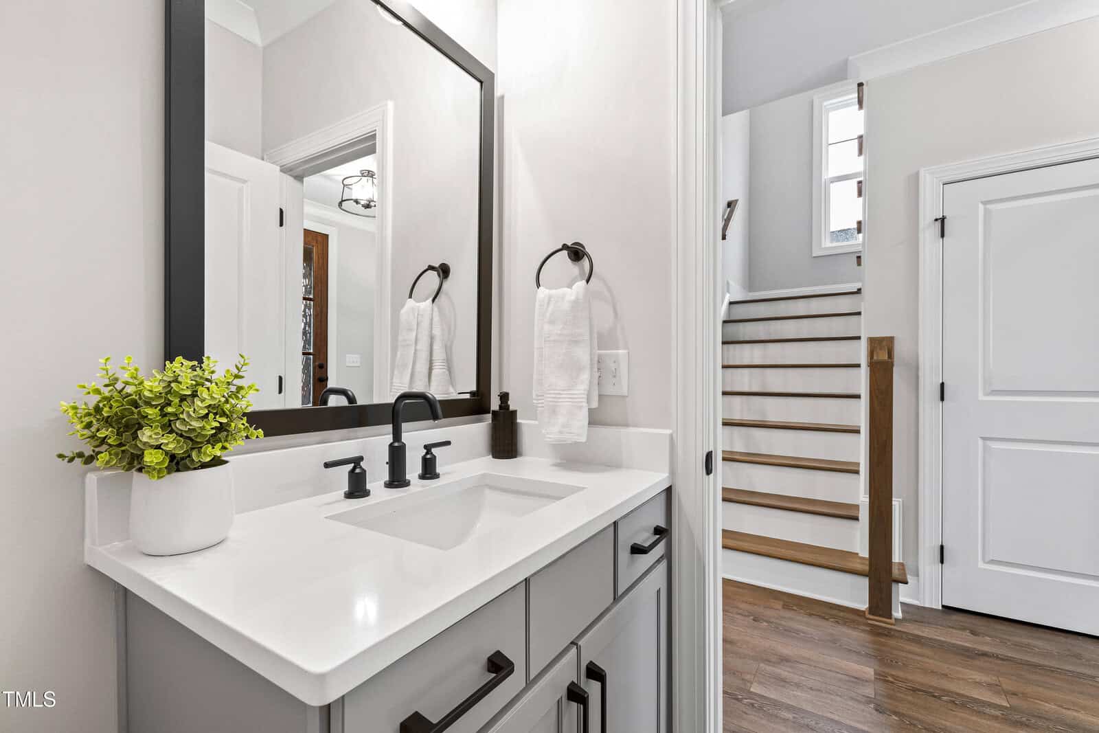 Modern bathroom with a white vanity, black fixtures, a large mirror, a potted plant, and neatly folded towels. In the background, there is a wooden staircase and a white door.