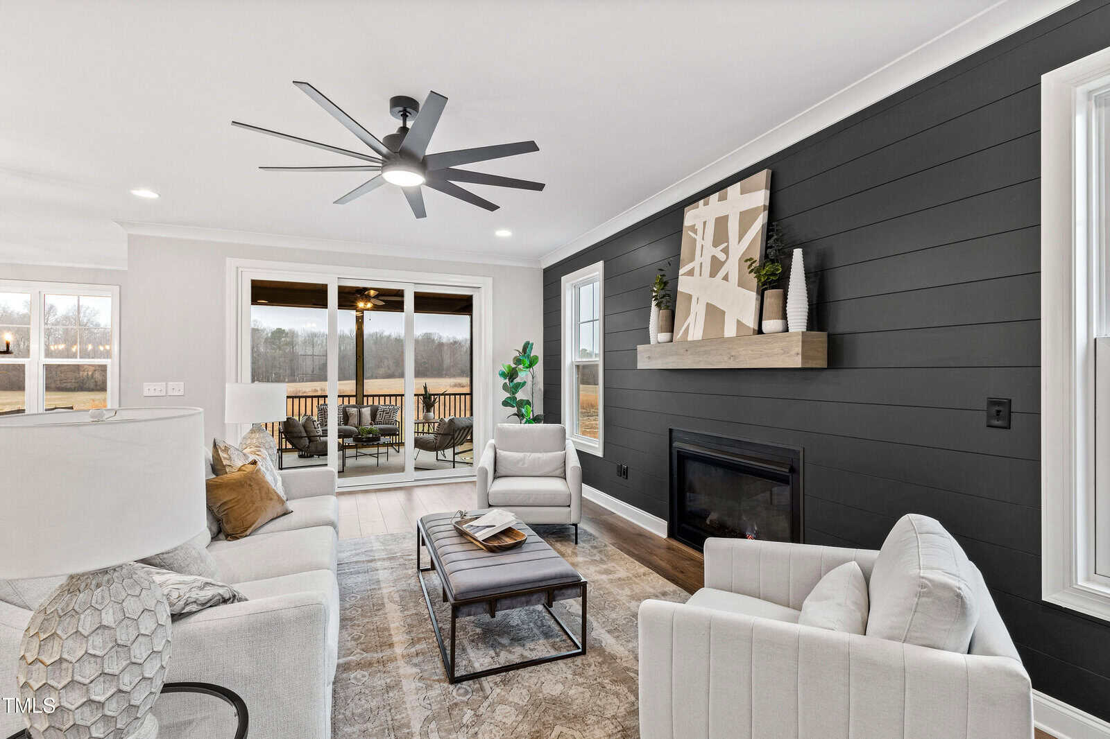 Modern living room with light-colored sofas and chairs, black accent wall with a fireplace, large abstract art above mantle, coffee table, ceiling fan, floor plant, and sliding glass doors opening to a deck with a scenic outdoor view.