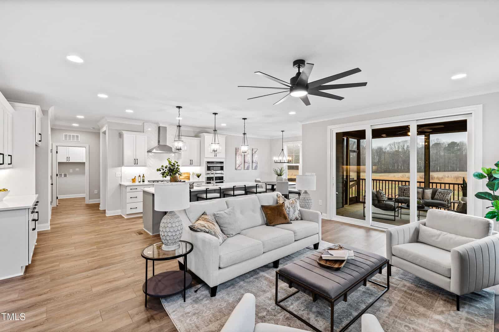 Modern open-concept living room and kitchen with white cabinets, stainless steel appliances, light wood floors, neutral furnishings, a large ceiling fan, and sliding glass doors leading to a screened porch with outdoor seating.