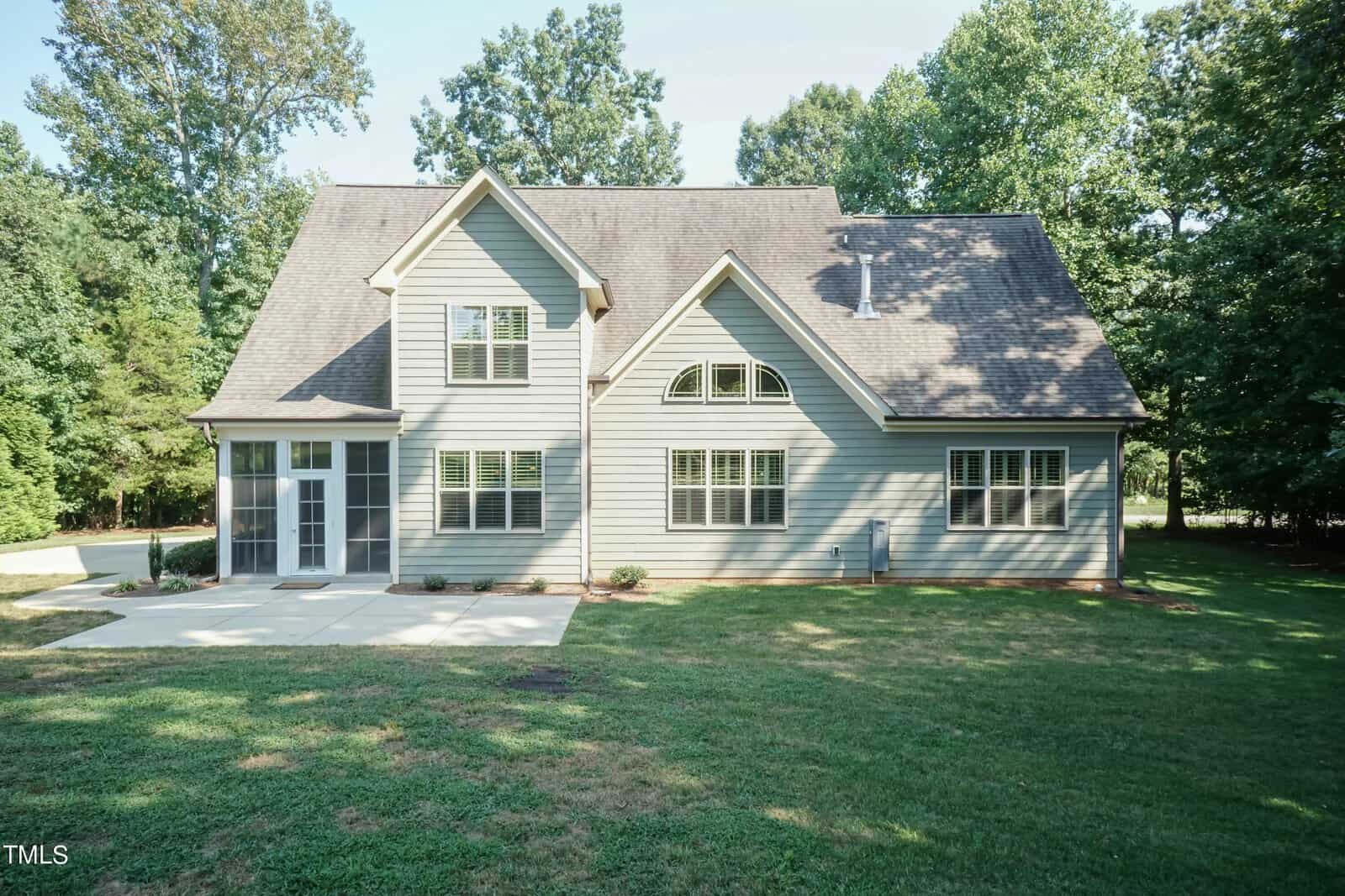 A two-story, light gray house with multiple windows and a gabled roof. It has a small front porch and is surrounded by a grassy yard with trees in the background, under a clear sky.