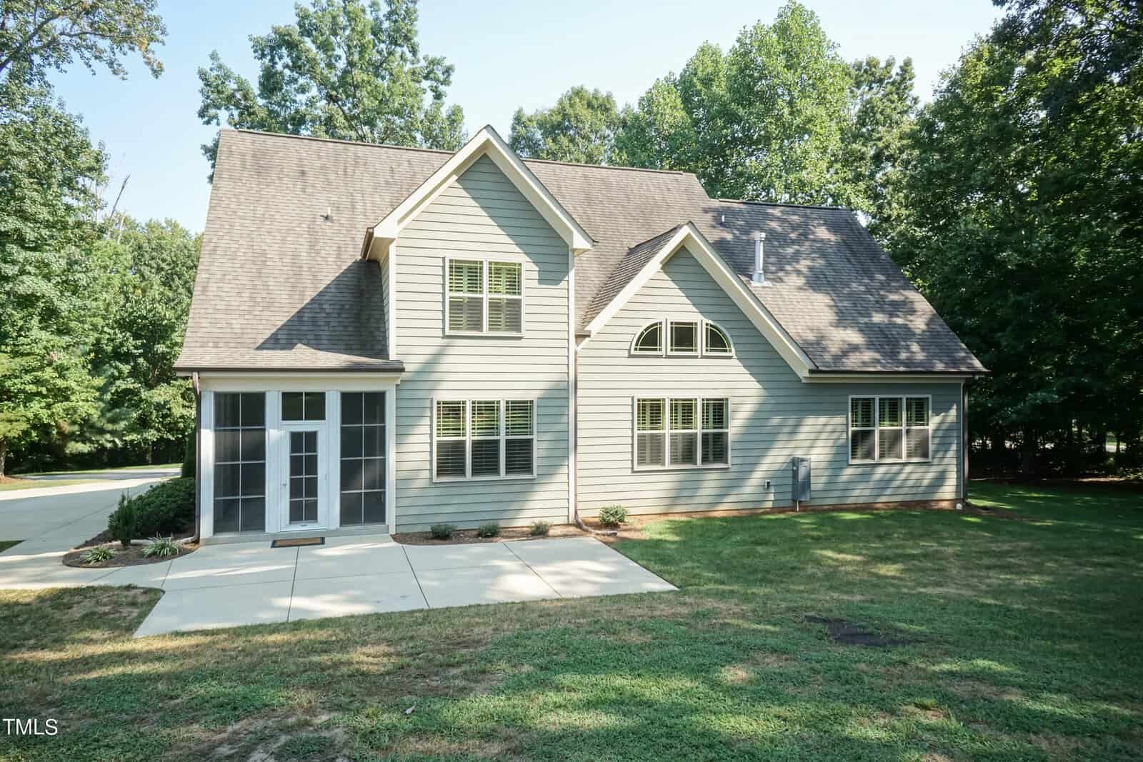 A modern two-story house with gray siding and multiple windows, including arched ones, set in a lush green yard with trees in the background. A concrete pathway leads to the entrance.
