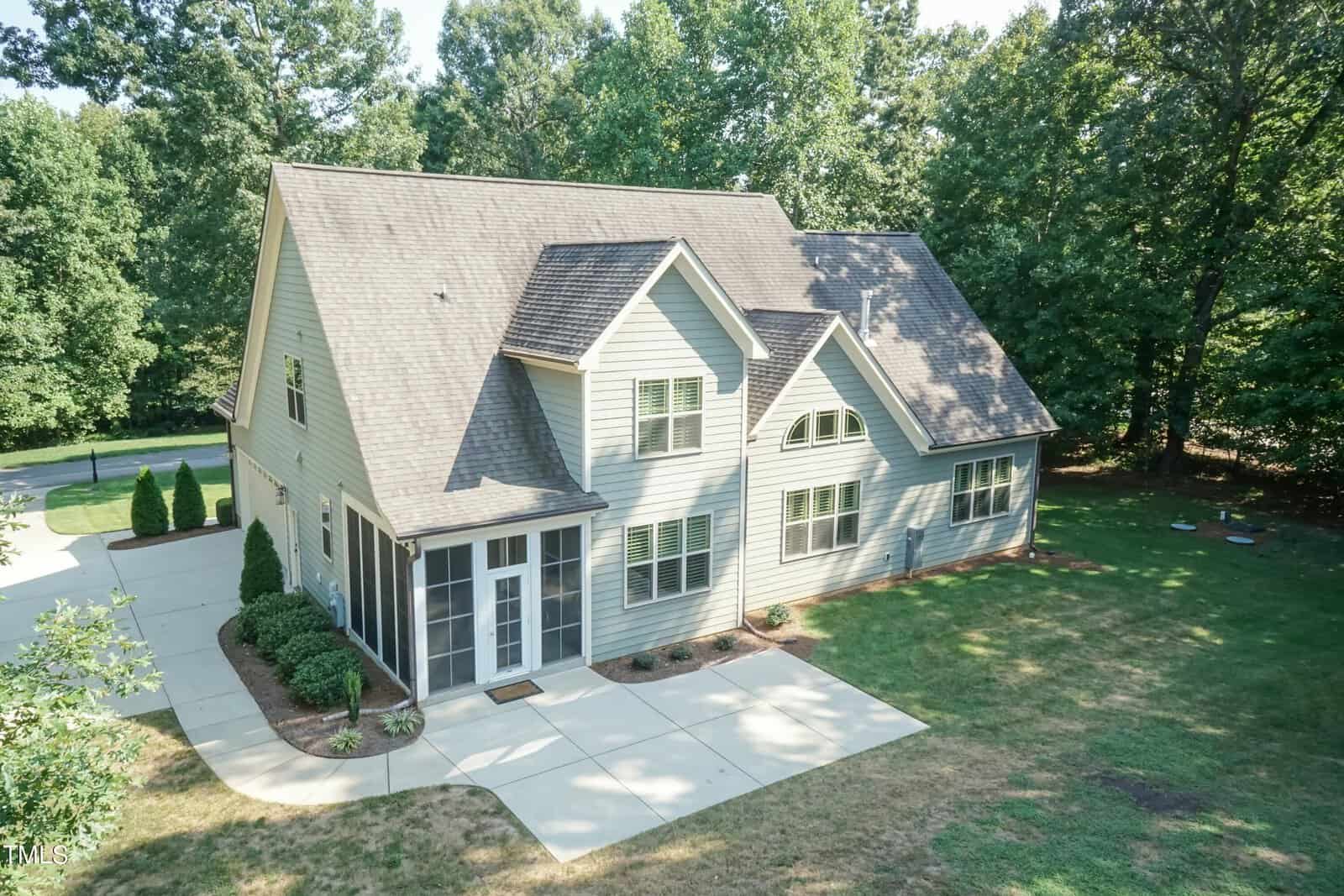 Aerial view of a two-story house with a pitched roof, light-colored siding, and a screened porch. The property is surrounded by trees and has a paved driveway and walkway, with neatly trimmed grass and minimal landscaping.