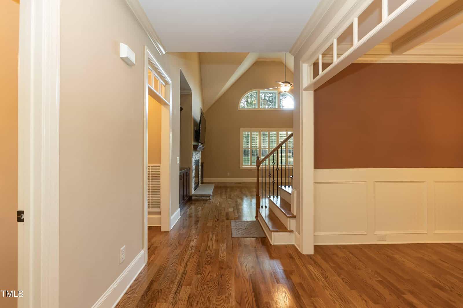 A bright interior hallway with hardwood floors and beige walls. A staircase with wooden railings is on the right, leading to a higher floor. An arch window and a ceiling fan are visible in a high-ceiling living room.