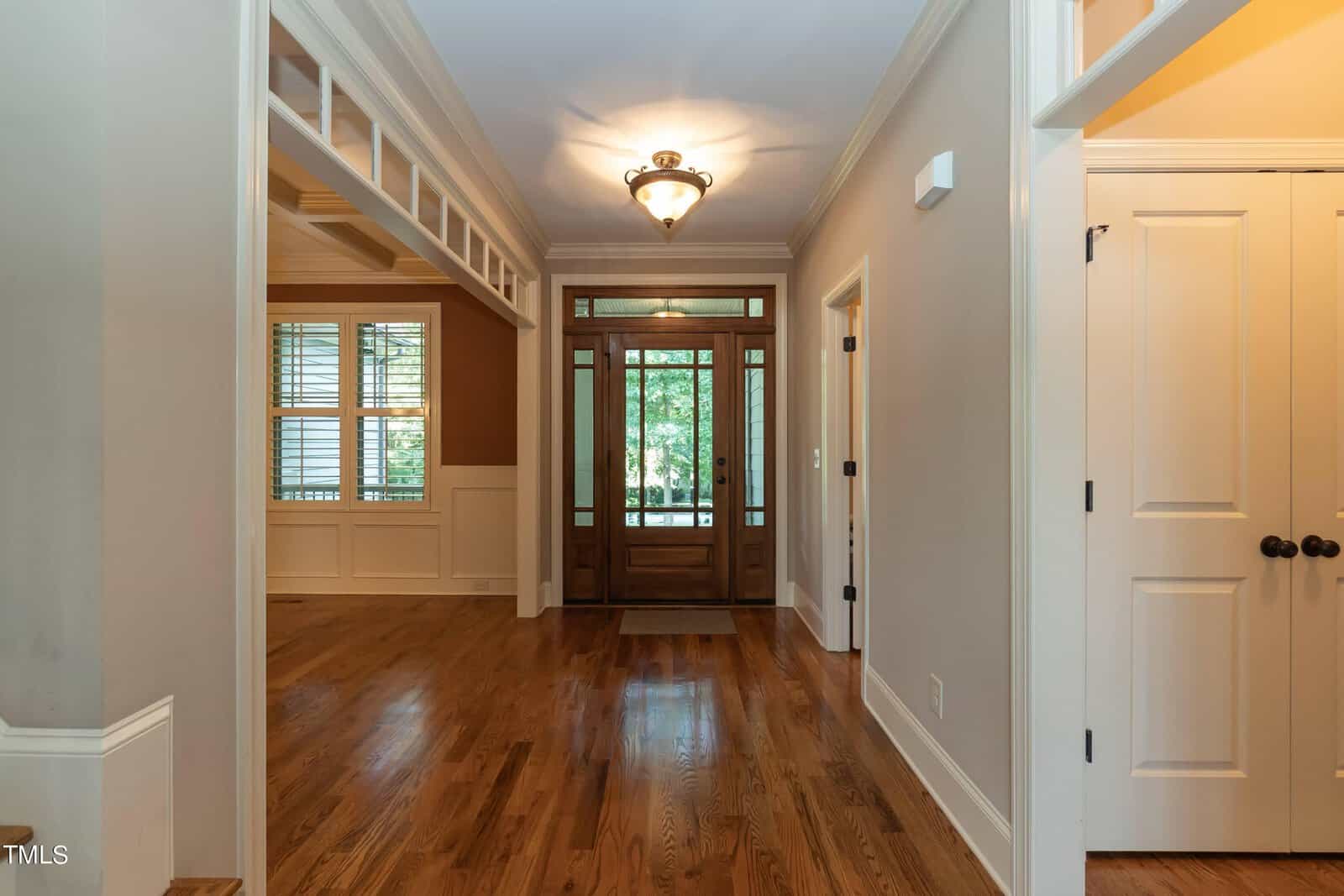 A spacious entryway with polished hardwood floors and a wooden front door featuring glass panels. To the left is a room with large windows and white wainscoting. The walls are painted in neutral tones, and a ceiling light fixture hangs in the hallway.